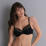 Žena s dlouhými hnědými vlasy má na sobě podprsenku Anita Colette 5248 s fialovou a zelenou květinovou krajkou, stojí před šedým pozadím