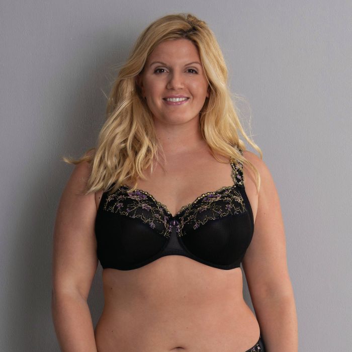 Žena s dlouhými blond vlasy, oblečená v černé podprsence Anita Colette 5249 s květinovou výšivkou, stojí před šedým pozadím a usmívá se do kamery.